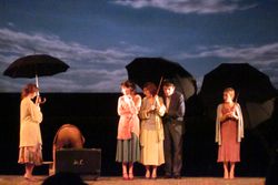 una imagen de una de las primeras escenas de la obra con todos los actores en escena ataviados con paraguas 