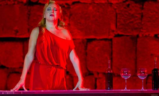carmen machi en un momento muy dramatico del monologo con todo el fondo rojo y ella vestida de rojo, rodeada de copas y botellas