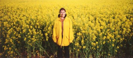 Isabel bono, brazos, piernas, cielo. Foto de ella en un campo de flores amarillas.